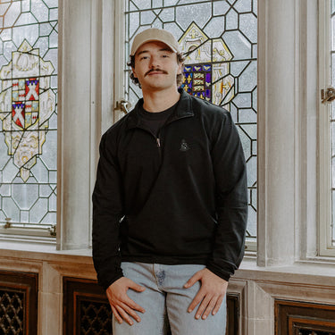 Male model wearing the black Alpha Sigma Phi Embroidered Luxe Quarter Zip in front of stained glass windows with fraternity crests.