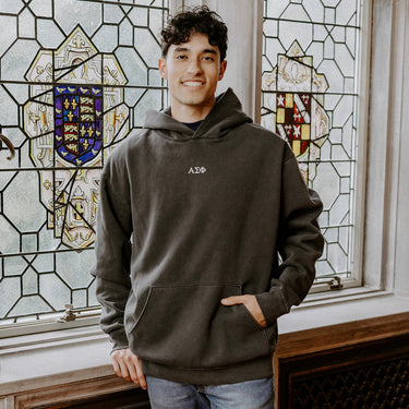 College-aged man wearing the Alpha Sigma Phi embroidered black hoodie, standing in front of stained glass windows with fraternity-style crest designs.
