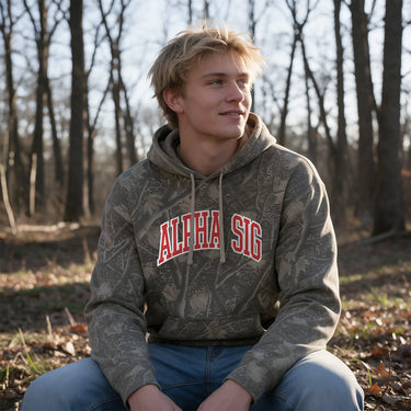 College-aged male wearing the fraternity Deadwood Camo Hoodie with red and white Alpha Sigma Phi lettering, seated outdoors in a wooded setting.