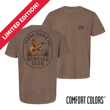 Brown garment-dyed short-sleeve tee featuring a vintage-inspired graphic: a weathered oval frames a mallard in mid-flight with burnt orange and mossy green-tipped wings above rippling water and cattails; distressed Alpha Sigma Phi arcs overhead and Alpha Sig letters bold and rugged on the front.